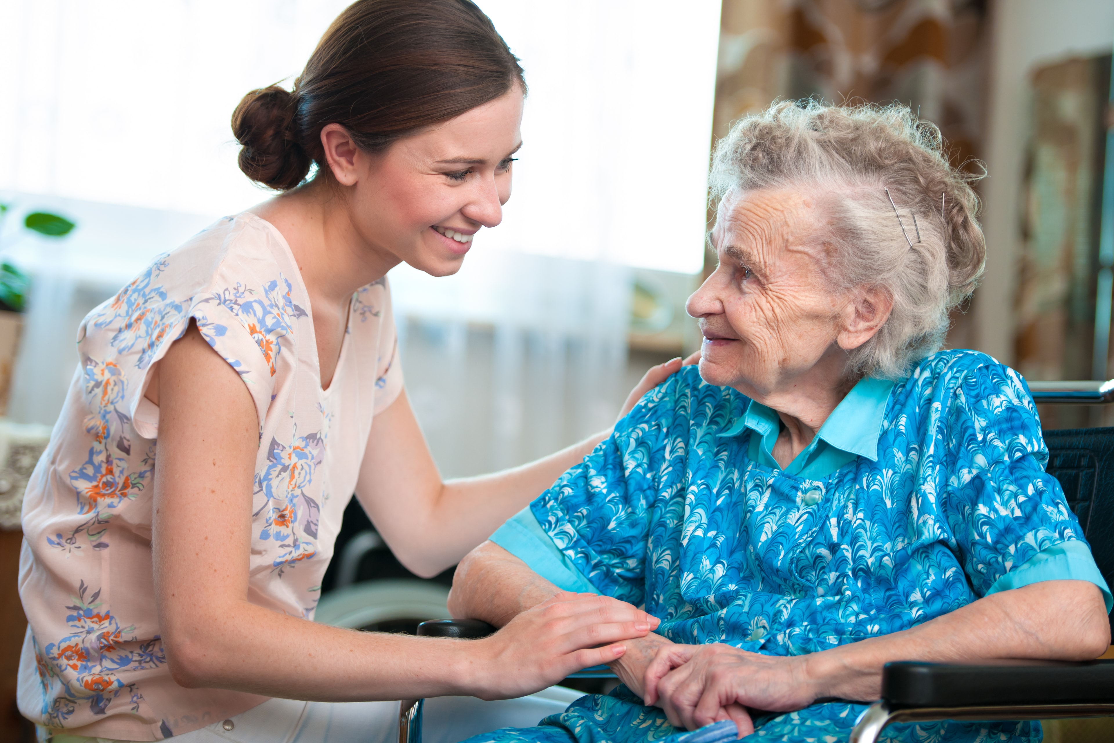 Young caregiver smiling warmly at an elderly woman in a wheelchair, holding her hands.